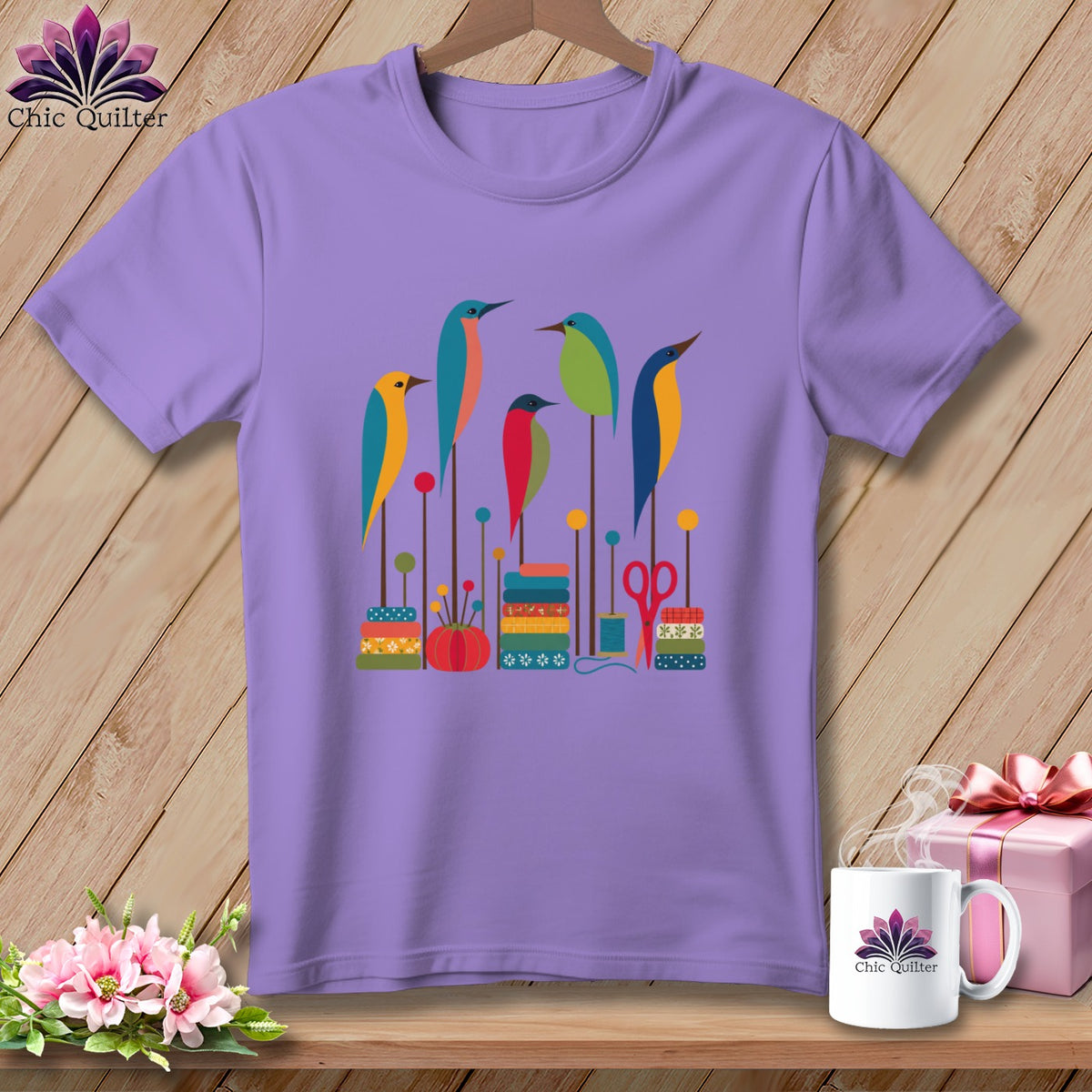 Lavender t-shirt featuring a colorful graphic of five stylized birds perched on sewing supplies against a wooden background.
