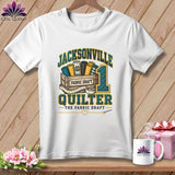 A white t-shirt featuring a Jacksonville Quilter graphic with fabric bolts, a wooden crate, and a measuring tape.