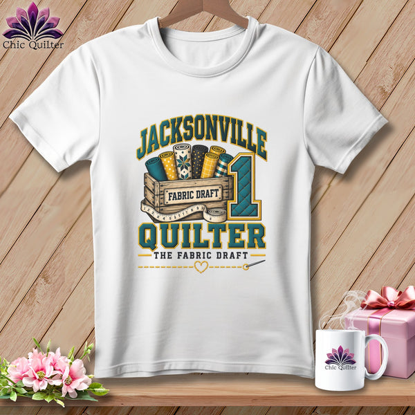 A white t-shirt featuring a Jacksonville Quilter graphic with fabric bolts, a wooden crate, and a measuring tape.