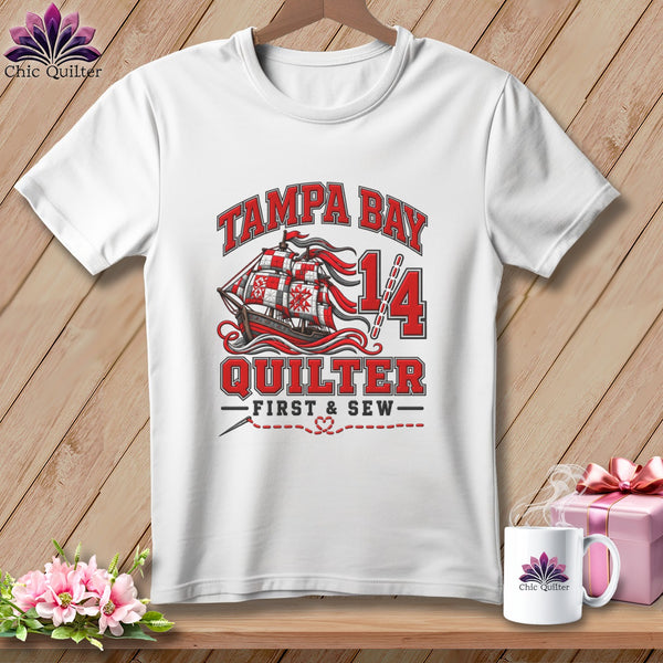 A white t-shirt hanging on a wooden background, featuring Tampa Bay Quilter graphics and a sailing ship.