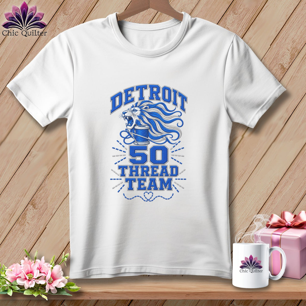 A white crewneck t-shirt hanging on a wooden background, featuring blue graphic text reading Detroit 50 Thread Team with a lion and spool illustration.