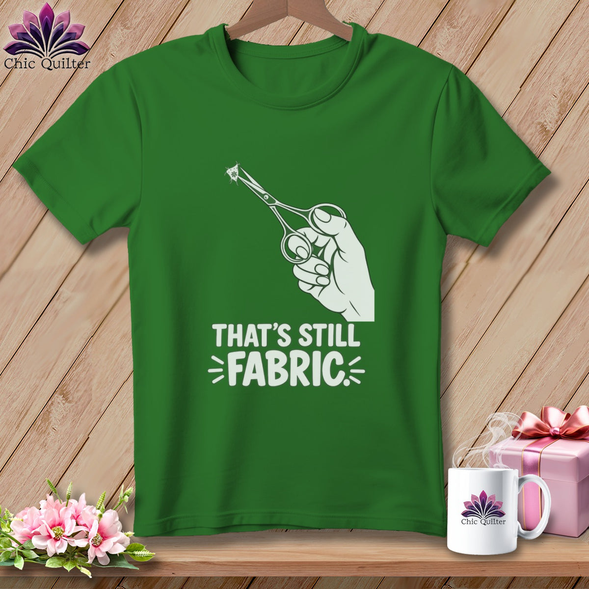 Green t-shirt with white graphic of hand holding scissors cutting fabric and text "THAT'S STILL FABRIC".