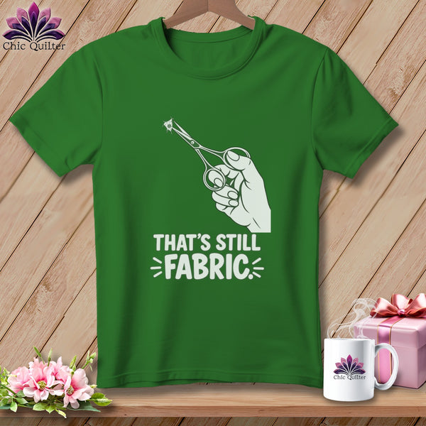 Green t-shirt with white graphic of hand holding scissors cutting fabric and text "THAT'S STILL FABRIC".
