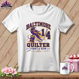 A white t-shirt hanging on a wooden background features a purple and yellow raven graphic with Baltimore Quilter First & Sew text.