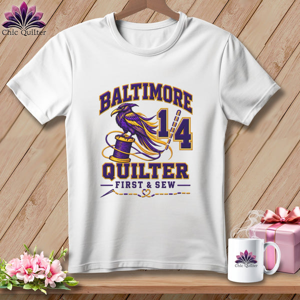 A white t-shirt hanging on a wooden background features a purple and yellow raven graphic with Baltimore Quilter First & Sew text.
