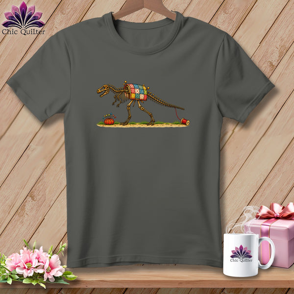 A dark grey t-shirt features a dinosaur skeleton graphic wearing a quilted blanket, accompanied by a pin cushion and thread spool.