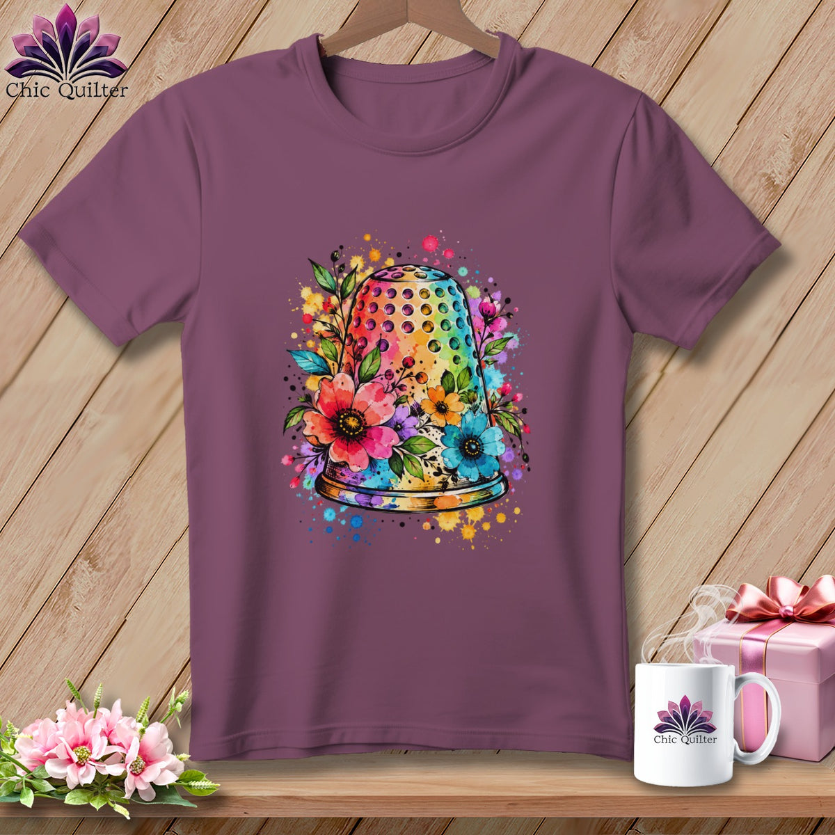 Mauve short-sleeve t-shirt featuring a watercolor thimble and floral graphic centered on the chest, displayed on a wooden background.