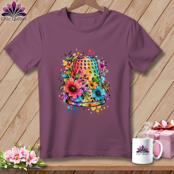 Mauve short-sleeve t-shirt featuring a watercolor thimble and floral graphic centered on the chest, displayed on a wooden background.