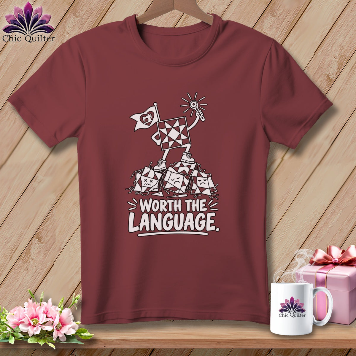 Maroon t-shirt with a graphic of a quilt square character holding a flag and a lightbulb over a pile of sad quilt squares with text "Worth the Language".