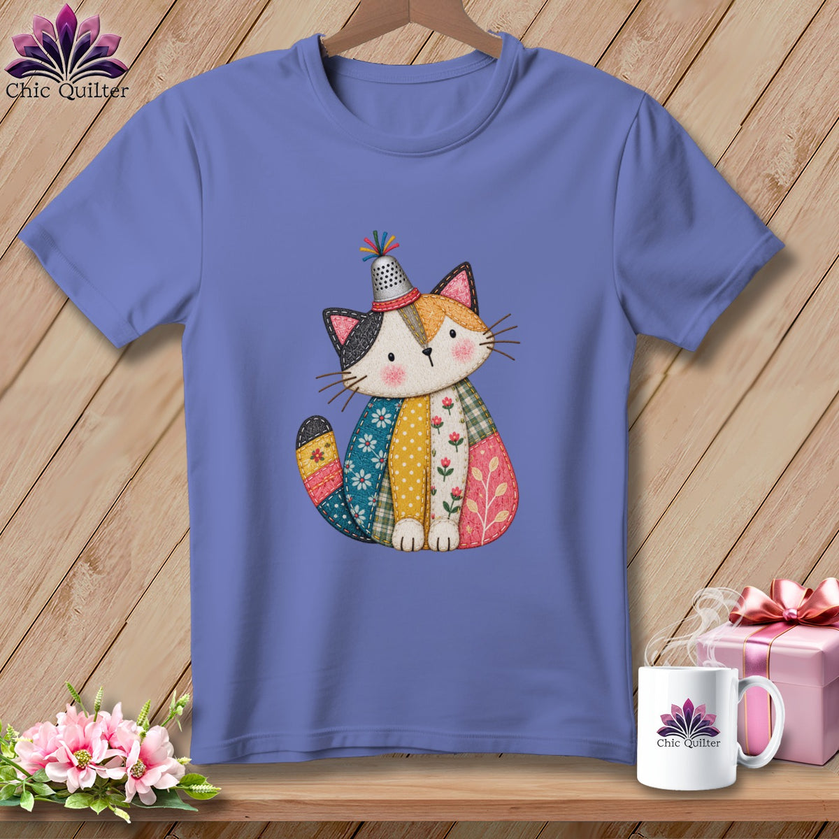 A periwinkle t-shirt features a graphic of a stitched patchwork cat wearing a thimble hat.