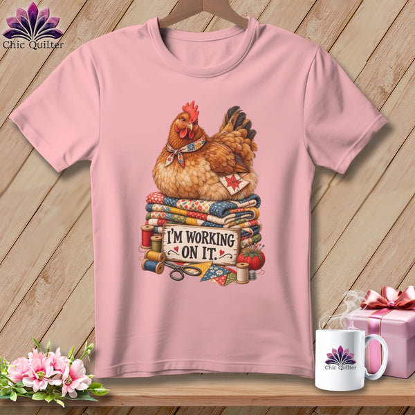 Pink t-shirt with a chicken on quilts and the text "I'M WORKING ON IT."