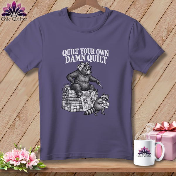 Purple t-shirt featuring a graphic of a bear and raccoon quilting with the text Quilt Your Own Damn Quilt.