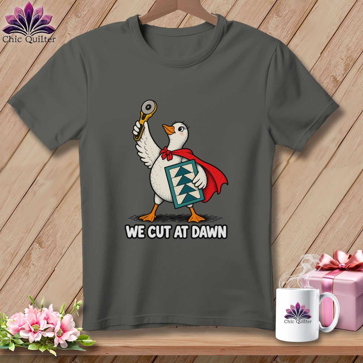 A dark gray t-shirt with a graphic of a goose wearing a red cape, holding a rotary cutter, and carrying a quilt block, with the text WE CUT AT DAWN below.
