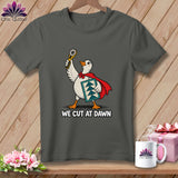 A dark gray t-shirt with a graphic of a goose wearing a red cape, holding a rotary cutter, and carrying a quilt block, with the text WE CUT AT DAWN below.