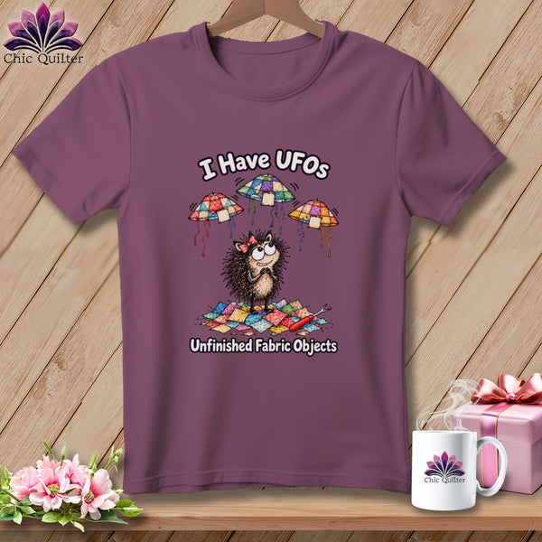 Mauve t-shirt featuring a cartoon hedgehog, flying quilted UFOs, and the text I Have UFOs Unfinished Fabric Objects.