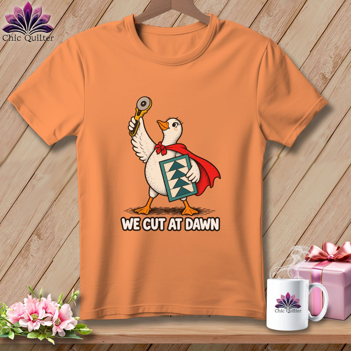 An orange t-shirt featuring a duck wearing a red cape, holding a rotary cutter and quilt block, with the text WE CUT AT DAWN.