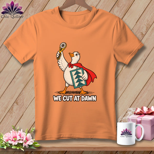 An orange t-shirt featuring a duck wearing a red cape, holding a rotary cutter and quilt block, with the text WE CUT AT DAWN.