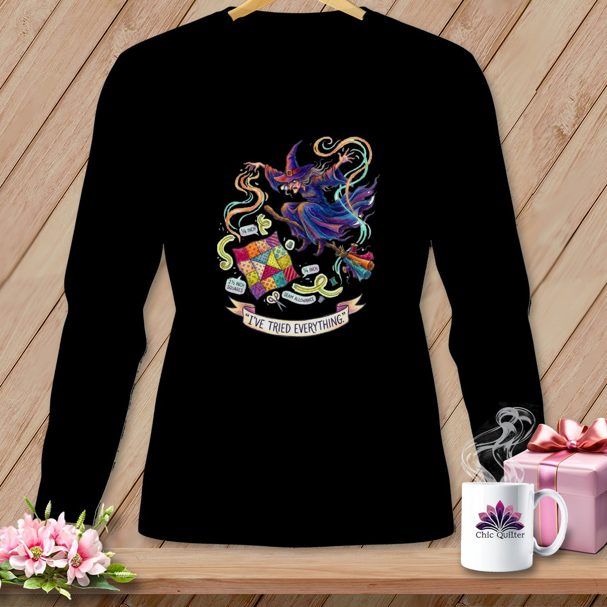 Black long-sleeve shirt with a colorful witch flying on a broomstick holding a patchwork quilt and text.