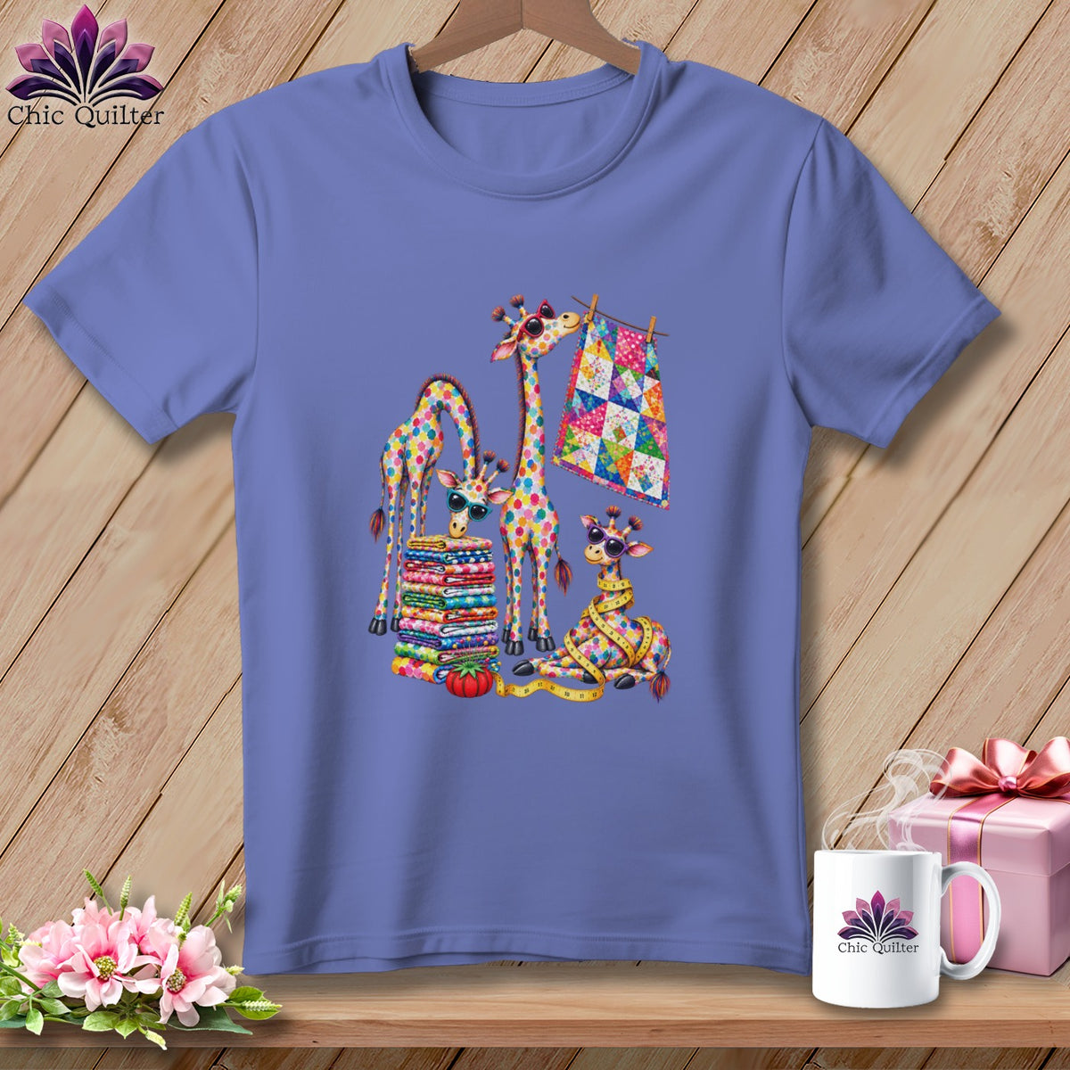 Periwinkle t-shirt featuring a colorful graphic of patchwork giraffes, a fabric stack, and a quilt on a wooden background.