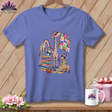 Periwinkle t-shirt featuring a colorful graphic of patchwork giraffes, a fabric stack, and a quilt on a wooden background.