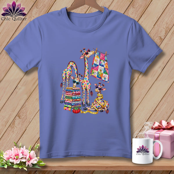 Periwinkle t-shirt featuring a colorful graphic of patchwork giraffes, a fabric stack, and a quilt on a wooden background.