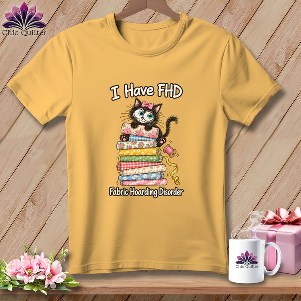 A mustard yellow t-shirt features a graphic of a black cat with a pink bow perched on a stack of colorful fabric rolls, with the text "I Have FHD Fabric Hoarding Disorder."