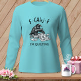 Turquoise long-sleeve shirt with a raven sewing a quilt, text reads 