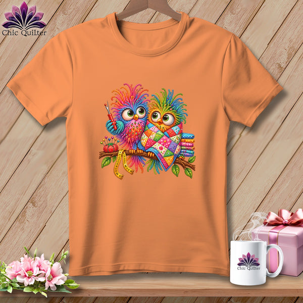 An orange short-sleeve t-shirt features a colorful graphic of two owls on a branch with quilting accessories.