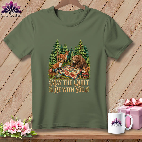 An olive green t-shirt displays a graphic of forest animals gathered around a colorful patchwork quilt with the text May The Quilt Be With You.