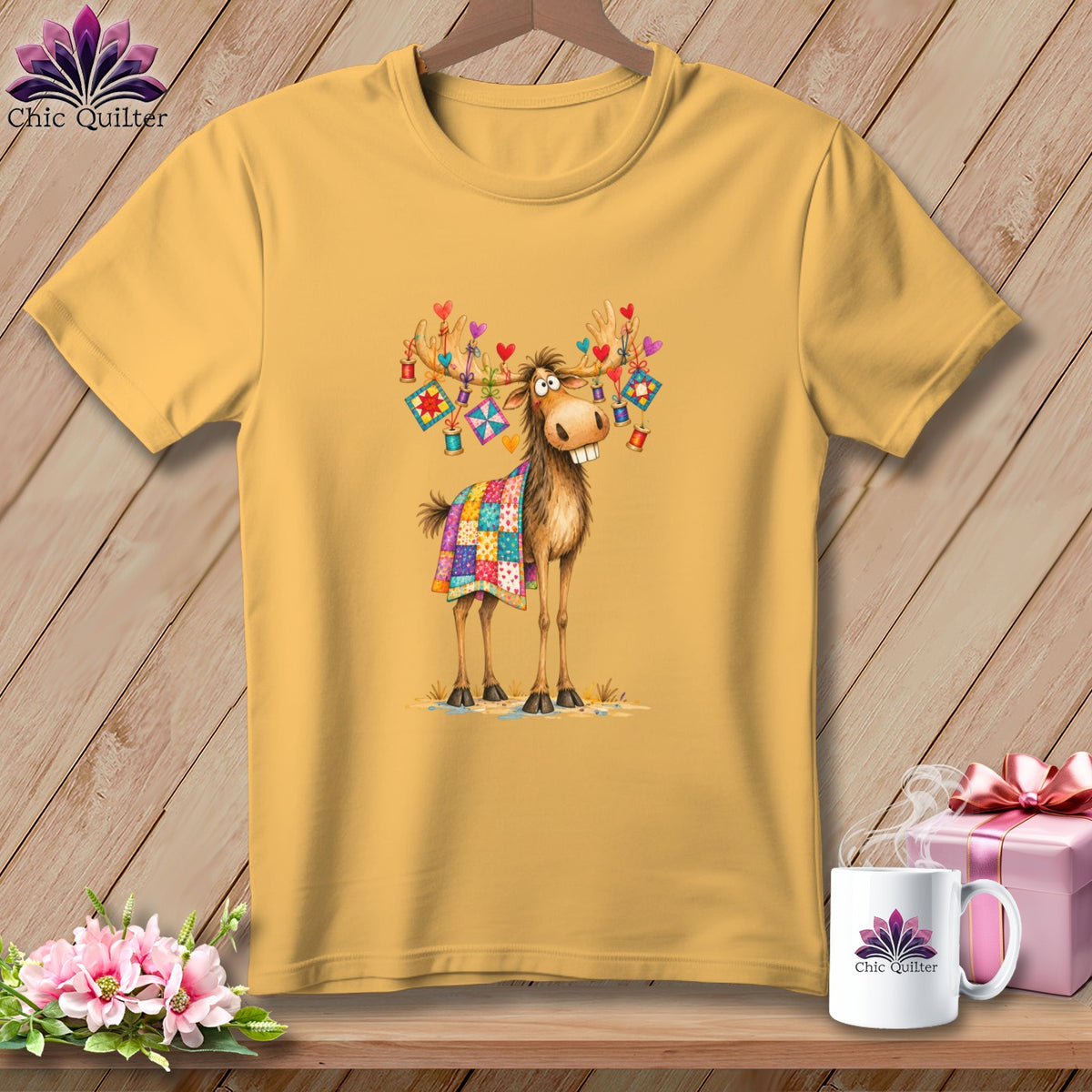 A yellow t-shirt featuring a cartoon moose draped in a colorful patchwork quilt and decorated antlers.