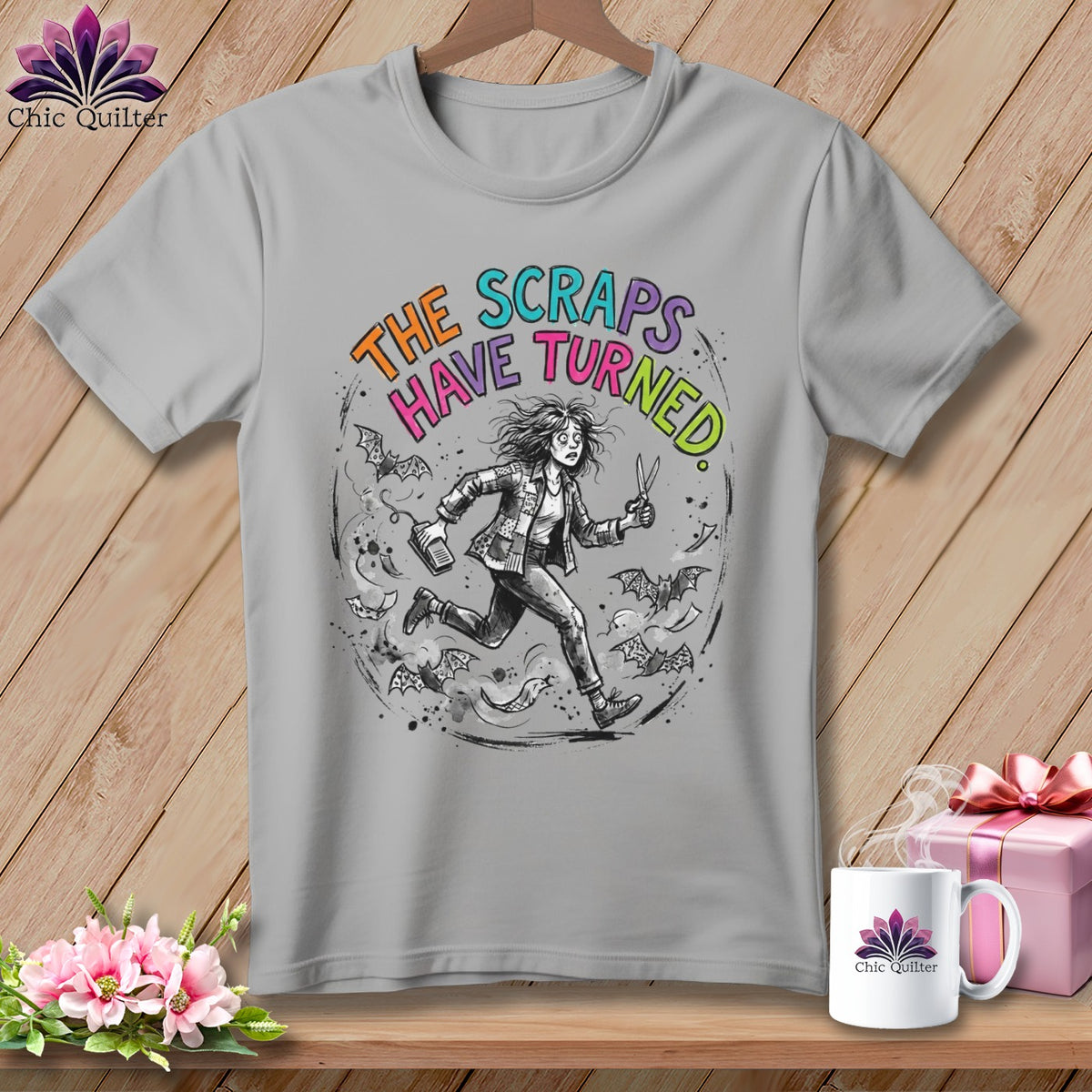 Gray t-shirt with illustration of a woman running with scissors and bats, text "THE SCRAPS HAVE TURNED".