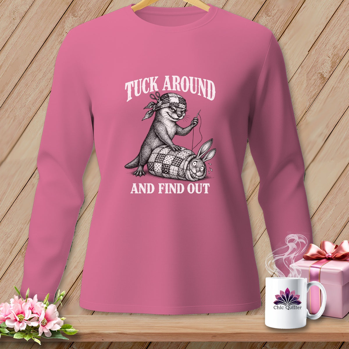 A pink long-sleeved shirt with a graphic of an otter sewing a quilt over a bound rabbit and text reading Tuck Around and Find Out.