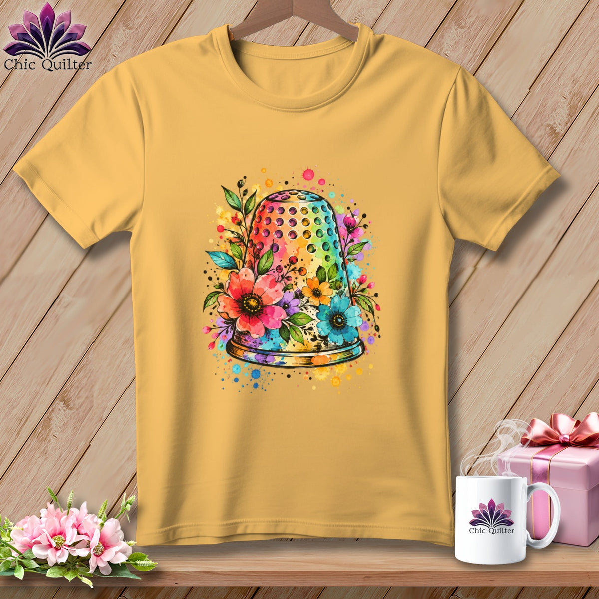 Yellow t-shirt hanging against a wooden wall, featuring a colorful watercolor graphic of a floral-decorated thimble.
