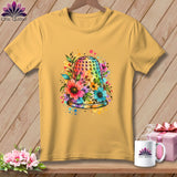 Yellow t-shirt hanging against a wooden wall, featuring a colorful watercolor graphic of a floral-decorated thimble.