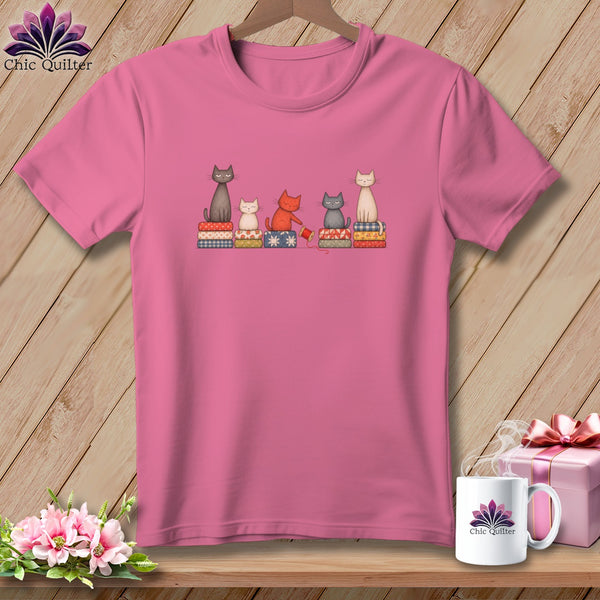 Pink t-shirt featuring an illustration of five cats sitting on quilted cushions, hanging against a wooden wall.