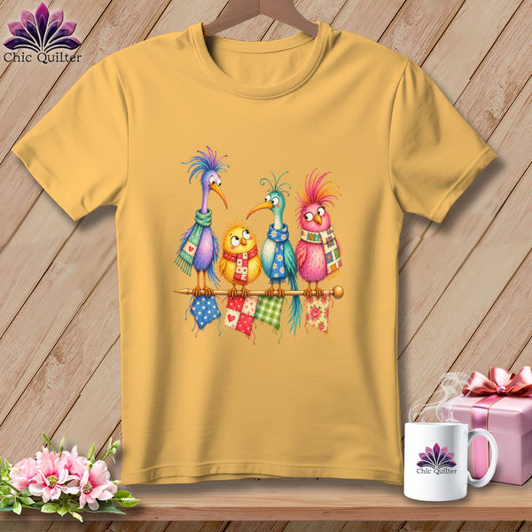 Gold t-shirt featuring four colorful cartoon birds wearing scarves while perched on a knitting needle.