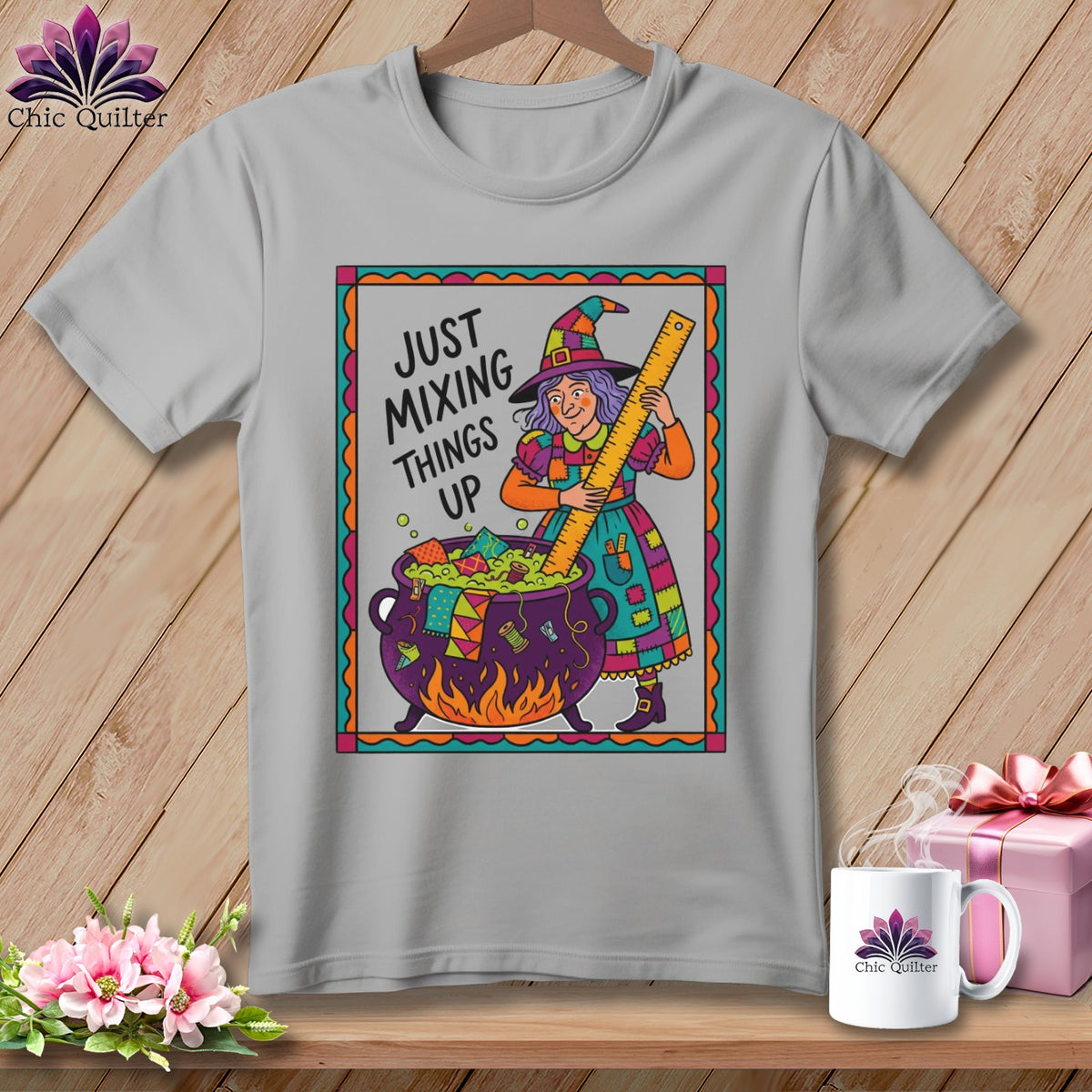 Gray t-shirt with a cartoon witch mixing colorful fabric scraps in a cauldron, text reads "Just Mixing Things Up".
