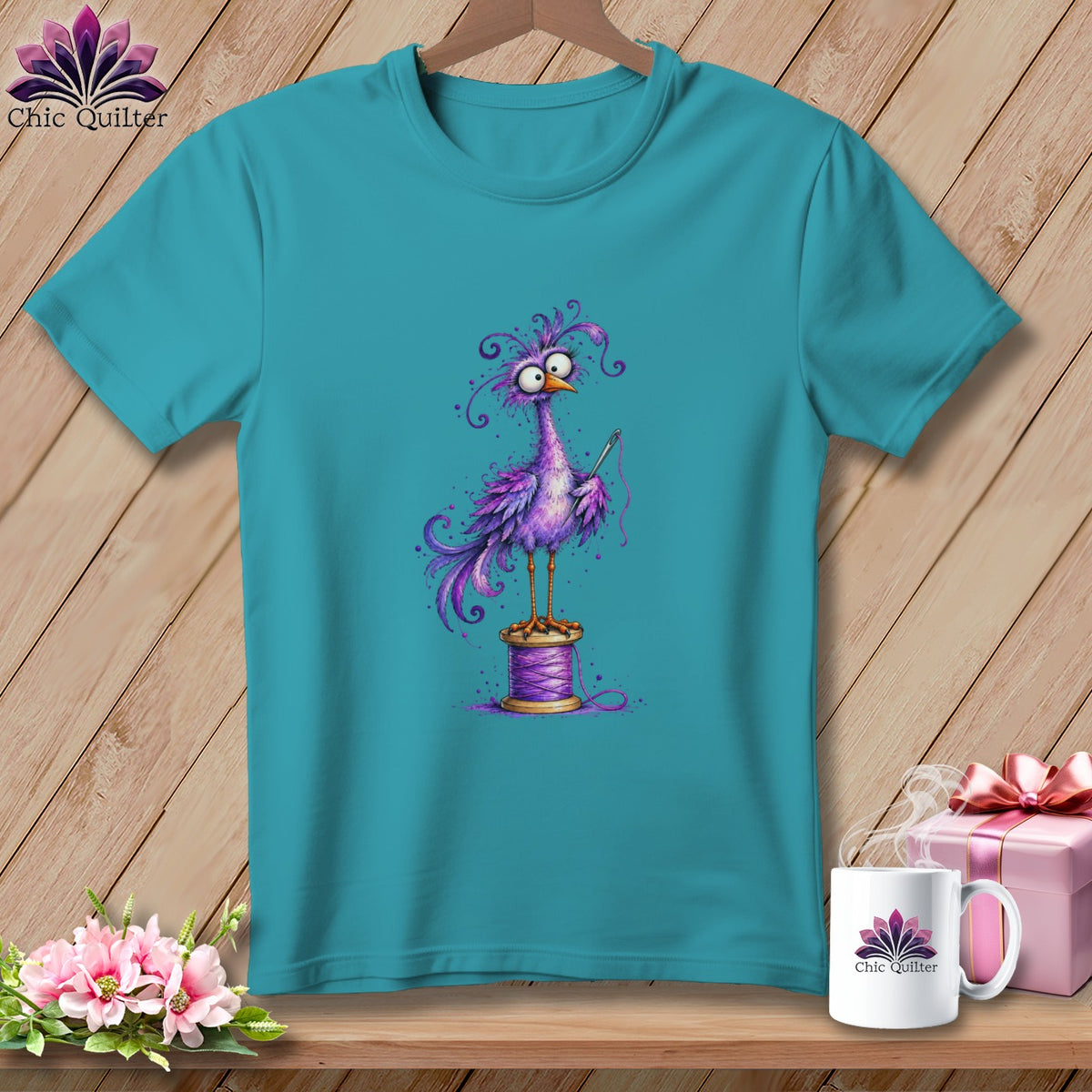 Teal t-shirt featuring a cartoon illustration of a quirky purple bird holding a sewing needle while standing on a spool of thread.