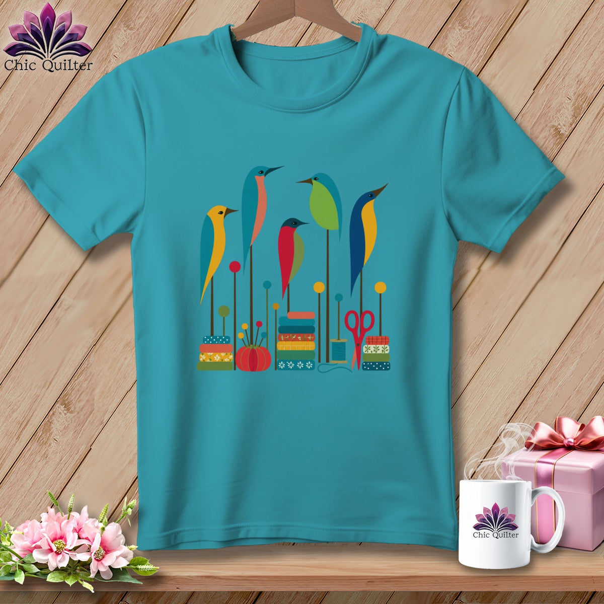 A teal crewneck t-shirt featuring a colorful graphic of stylized birds, spools, and sewing notions.