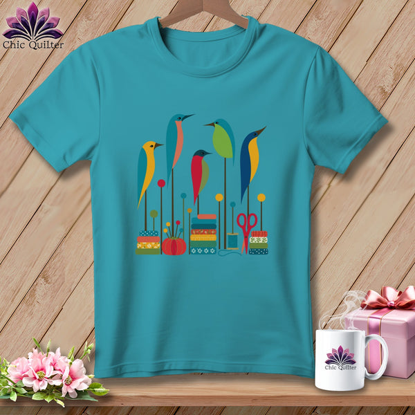 A teal crewneck t-shirt featuring a colorful graphic of stylized birds, spools, and sewing notions.
