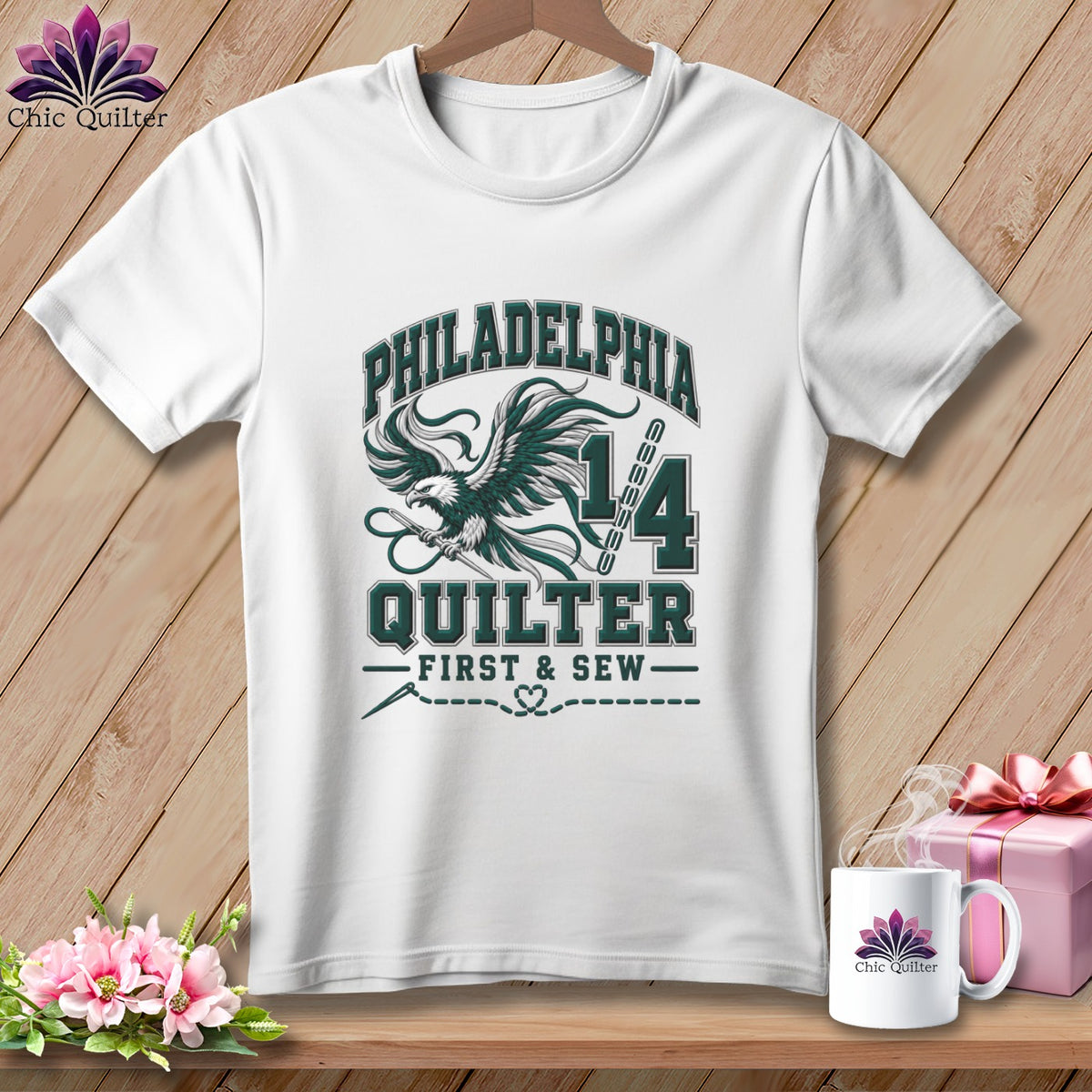 A white t-shirt hanging on a wooden background features a graphic with an eagle holding a needle, the text "PHILADELPHIA QUILTER FIRST & SEW," and the number 14.