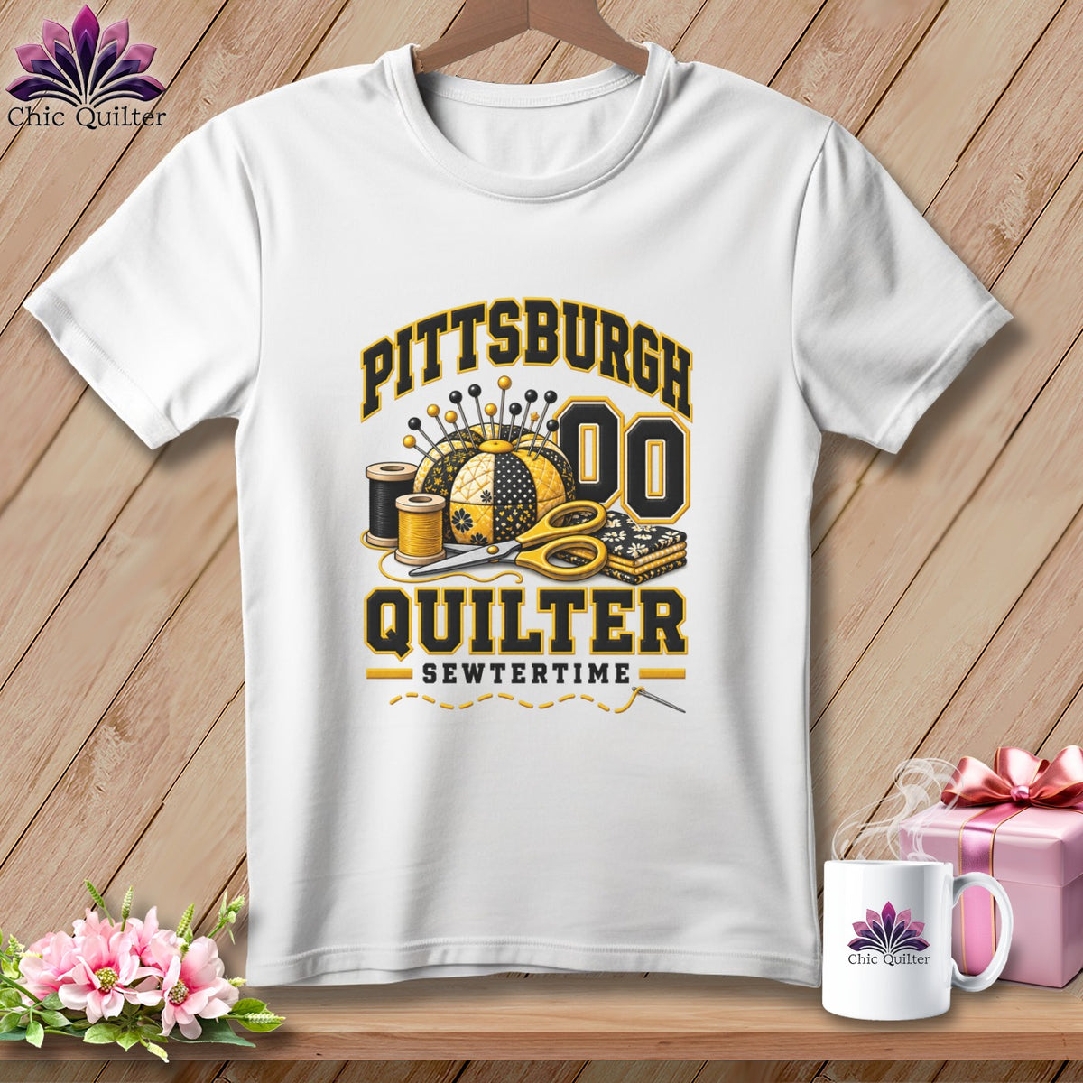 White t-shirt with a black and gold Pittsburgh Quilter graphic featuring sewing supplies against a wooden background.