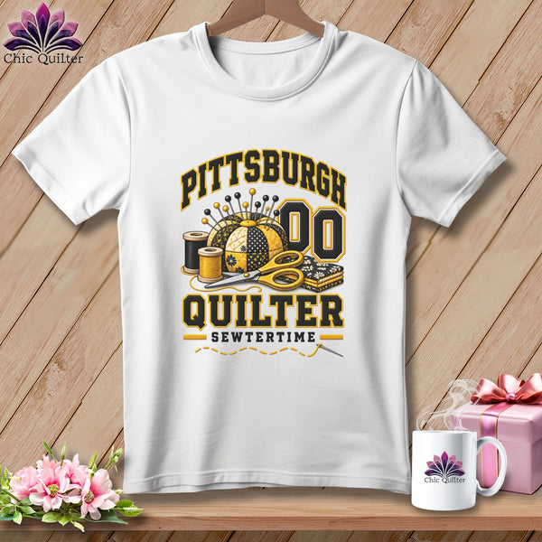 White t-shirt with a black and gold Pittsburgh Quilter graphic featuring sewing supplies against a wooden background.