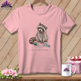 Pink t-shirt with raccoon, spool of thread, fabric, and text 