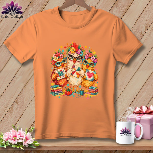 An orange t-shirt displays a graphic of three chickens wearing glasses and floral crowns while holding quilted blocks.