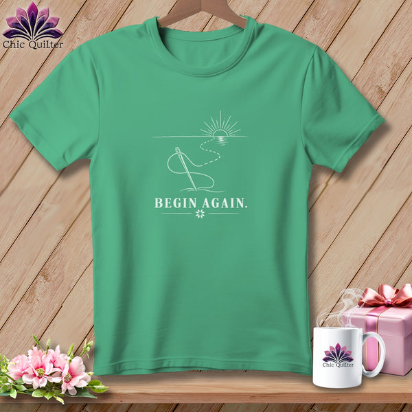Teal t-shirt with white line art of a needle, thread, sun, and water, reading "BEGIN AGAIN."