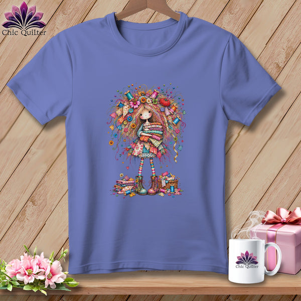 A periwinkle t-shirt features a colorful graphic of a girl holding folded quilts, surrounded by sewing accessories.
