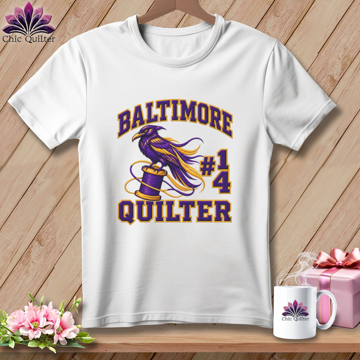 A white t-shirt hanging on a wooden background features a purple and gold raven graphic on a spool of thread with text that reads Baltimore Quilter.