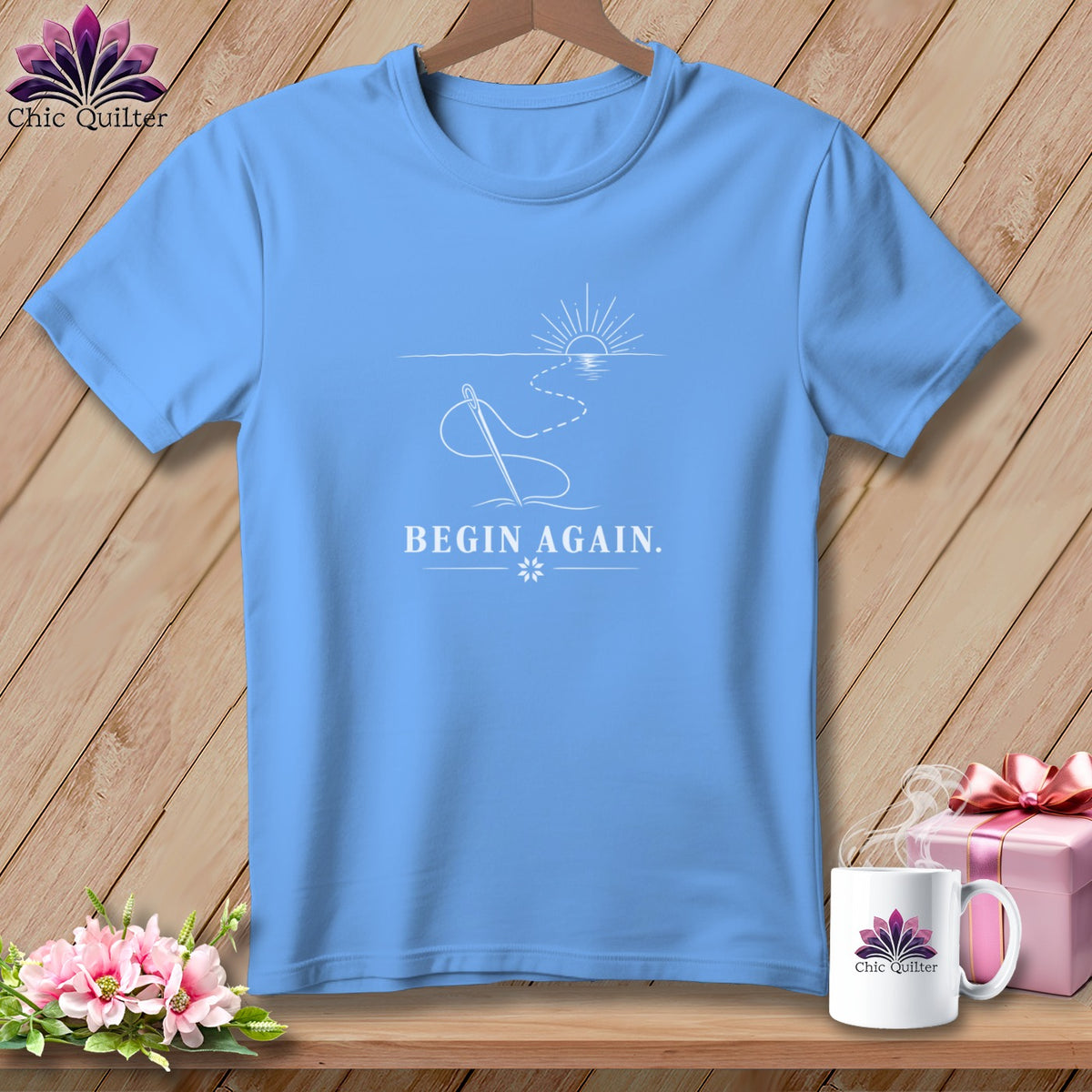 Blue t-shirt with a white line drawing of a needle, thread, sun, and the text "BEGIN AGAIN."