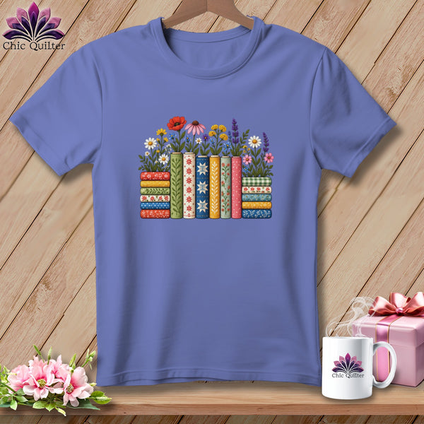 A periwinkle t-shirt features a graphic of colorful books stacked horizontally and vertically with flowers blooming from them.